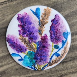 May include: A round, white coaster with a painted design of purple, blue, and gold flowers. The flowers are arranged in a bouquet and have a watercolor effect.