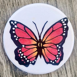 May include: A circular white object featuring a central illustration of a butterfly. The butterfly has wings with a gradient of red, orange, and yellow, outlined in black with white spots along the edges and two thin black antennae extending upward.