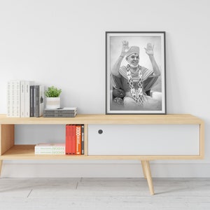 May include: A black and white photo of a man wearing a white turban and a long white robe with a beaded necklace. The photo is framed and hanging on a white wall above a wooden shelf with books and a potted plant.
