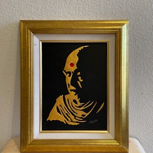 May include: A framed portrait of a person with a gold silhouette on a black background. The person has a red dot on their forehead.