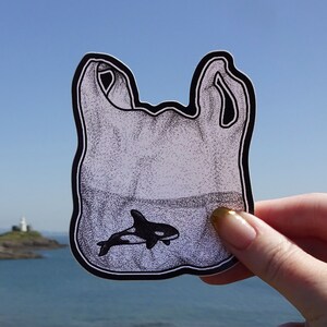 May include: A black and white illustration of a plastic bag with a small orca whale swimming inside. The bag is held in front of a blue sky and a body of water with a lighthouse in the distance.