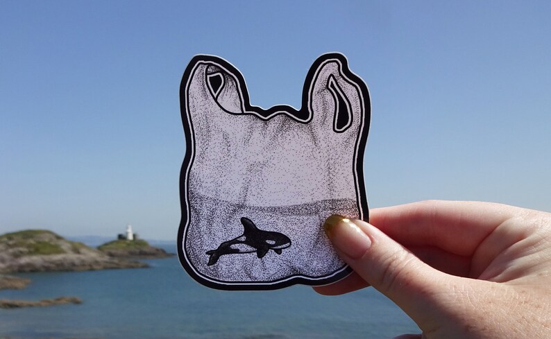 May include: A black and white illustration of a plastic bag with a small orca whale swimming inside. The bag is held in front of a blue sky and a body of water with a lighthouse in the distance.