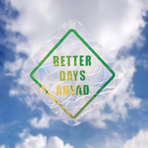 以下が含まれることがあります： 緑と黄色の「BETTER DAYS AHEAD」という文字が入ったひし形のステッカー。ステッカーは、テクスチャ加工された虹色の表面と緑色の縁取りがあり、白い雲のある青い空を背景にしています。