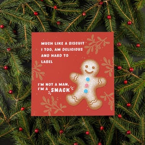 May include: A red greeting card featuring a gingerbread man illustration and the text "MUCH LIKE A BISCUIT I TOO, AM DELICIOUS AND HARD TO LABEL I'M NOT A MAN, I'M A SNACK". The card is decorated with sprigs and is surrounded by evergreen branches.