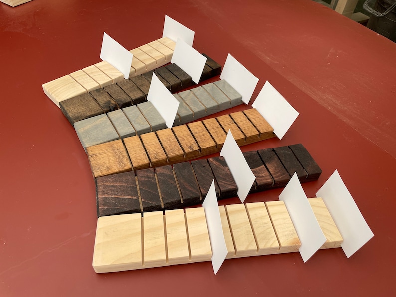 May include: A set of eight wooden business card holders in various wood stains, including natural, light brown, gray, dark brown, and black. Each holder has eight slots for business cards and is displayed with blank white cards.