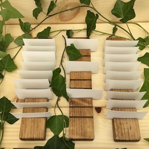 May include: Three wooden place card holders with white card inserts. Each holder has a rustic brown finish and is made of wood. The holders are arranged in a row on a light wood surface.