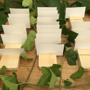 May include: A set of wooden place card holders with blank white cards. The holders are made of natural wood and have a rustic look. The cards are perfect for displaying names or messages at a wedding, party, or other event.