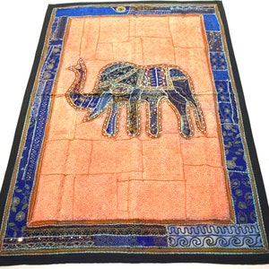 Puede incluir: Un tapiz de pared con un diseño de patchwork que presenta un elefante bordado en tonos azules, morados y dorados sobre un fondo melocotón. El borde es una mezcla de azul, dorado y negro.