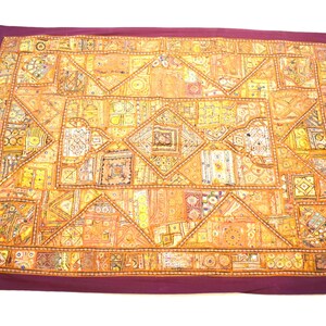 May include: A large, rectangular tapestry with a patchwork design. The tapestry is made of various fabrics in shades of gold, brown, and white. The tapestry is surrounded by a dark purple border.
