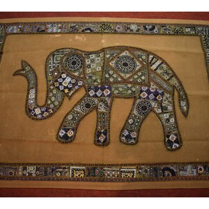 Puede incluir: Un tapiz de color beige con un gran elefante bordado en el centro. El elefante está hecho de varios retazos de tela y lentejuelas en tonos dorados, plateados, morados y negros.