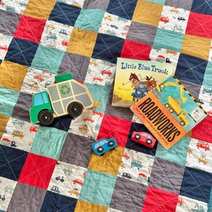 May include: A patchwork quilt in squares of red, blue, yellow, and grey, decorated with car patterns. Wooden toy cars and a green and grey wooden toy refuse truck are on the quilt, alongside two children's books.
