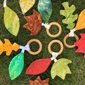 May include: A handmade baby toy with wooden rings and fabric leaves in various colors and patterns, including green, red, orange, and brown. The leaves are sewn together with white ribbon.