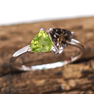 May include: A silver ring with two triangular gemstones, one light green and one smoky brown. The ring is displayed against a natural wood background, emphasising the gemstones' colours and the ring's design.