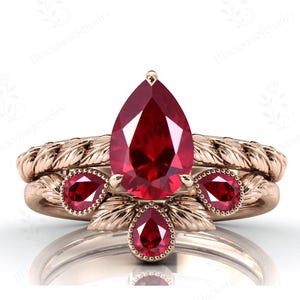 Peut inclure: Ensemble de bagues en or rose avec une pierre centrale en rubis en forme de poire et de petits accents de rubis. La bande présente un motif tressé. La bague est présentée sur fond blanc.