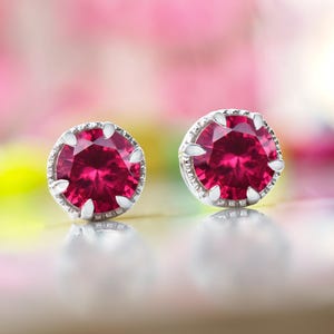 Puede incluir: Un par de pendientes de botón con piedras preciosas redondas de color rojo intenso engastadas en metal plateado. Las piedras preciosas son el punto focal, con una apariencia facetada. Los pendientes se exhiben sobre una superficie reflectante.