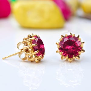 以下が含まれることがあります： 大きなルビーレッドの宝石が付いた、金色のスタッドピアス。ピアスは、詳細な透かし細工のデザインにセットされています。背景には、カラフルでぼやけた装飾用の石があります。