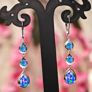 Puede incluir: Pendientes de plata con tres piedras de ópalo azul en forma de lágrima. Los pendientes se exhiben en un soporte negro sobre un fondo floral rosa borroso, resaltando los intrincados detalles de la joyería.