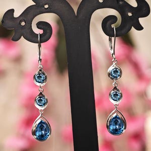 Può includere: Eleganti orecchini in argento con tre pietre blu ciascuno. Le due pietre superiori sono rotonde, mentre quella inferiore è a goccia. Gli orecchini pendono da ganci d'argento. Sfondo sfocato con fiori rosa.