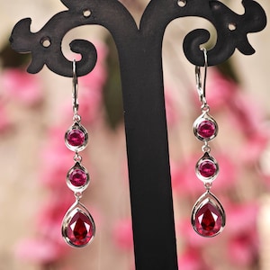 May include: Elegant silver drop earrings with three vibrant red gemstones. The earrings feature a teardrop-shaped stone at the bottom and two smaller round stones above. The jewelry is displayed on a black stand against a blurred pink background.