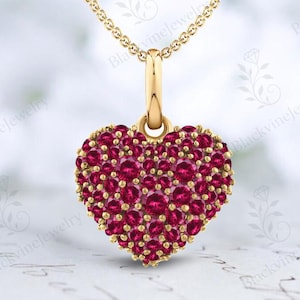May include: A gold pendant necklace featuring a heart-shaped charm. The charm is covered in small, round, red gemstones. The necklace chain and the charm's bail are also gold. The background is a soft, blurred white.