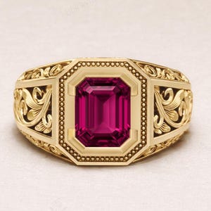 May include: A gold-colored ring with an emerald-cut, deep pink gemstone at the center. The ring has an ornate, engraved design on the band and around the gemstone setting. The ring is a statement piece of jewelry.