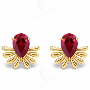 May include: A pair of gold-colored stud earrings. Each earring features a teardrop-shaped, deep red gemstone set atop a stylized, sunburst design. The earrings are displayed against a white background.