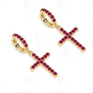 May include: A pair of gold-colored hoop earrings with dangling cross pendants. Each earring features a hoop lined with small, red gemstones and a cross pendant also adorned with red gemstones. The earrings are displayed against a white background.