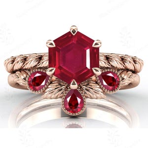 Puede incluir: Un juego de anillos de oro rosa con una gran piedra central de rubí hexagonal. El conjunto incluye una banda a juego con pequeños detalles de rubí y un diseño trenzado. El diseño del anillo incorpora detalles en forma de hojas.