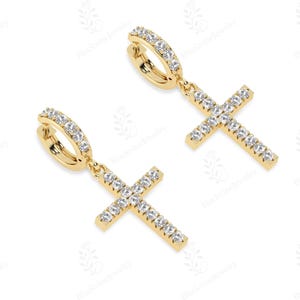 May include: A pair of gold-colored hoop earrings with dangling cross pendants. Each earring features a hoop lined with small, clear gemstones. The cross pendants are also adorned with similar gemstones, creating a sparkling effect.
