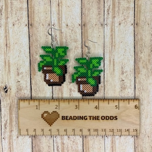 May include: A pair of green and brown pixelated plant earrings with silver hooks. The earrings are about 2 inches long and are displayed on a wooden ruler with the text "BEADING THE ODDS".