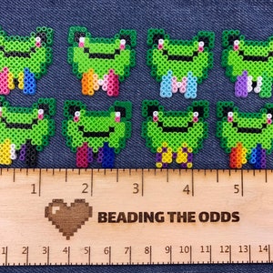 Puede incluir: Ocho sprites de cuentas en forma de rana verde con colas de arcoíris. Los sprites están dispuestos en dos filas de cuatro. Una regla de madera con el texto "BEADING THE ODDS" está debajo de los sprites.