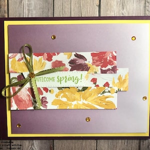 May include: A purple card with a yellow border and a floral design. The card has a green ribbon tied around it and the words "Welcome Spring!" written on it.