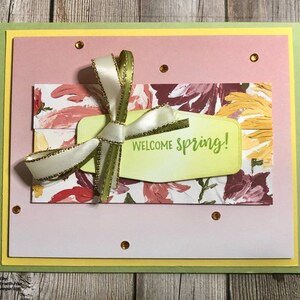 May include: A handmade greeting card with a pink and yellow floral design. The card features a green tag with the text "Welcome Spring!" and a white ribbon with gold trim.