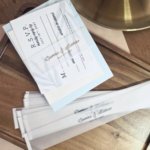 May include: Wedding invitation suite featuring a stack of clear vellum sleeves with the names "Laurena & Matthew" and the date "July 15th, 2027". The suite includes RSVP cards with a light blue floral design.