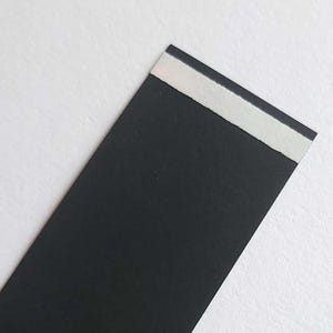 May include: A rectangular strip of matte black paper featuring a thin, horizontal white adhesive strip along the top edge. The paper has a smooth, solid black surface and a clean, straight-cut edge at the top where the white adhesive is applied.