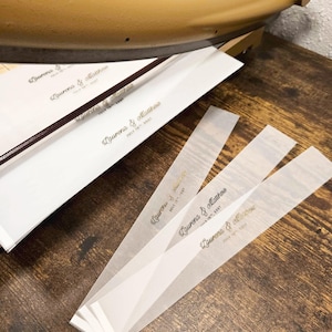 May include: Close-up of wedding invitation strips with the names "Lauren & Matthew" and the date "July 15th, 2023" printed in gold. The strips are on a wooden surface, with a paper cutter in the background.