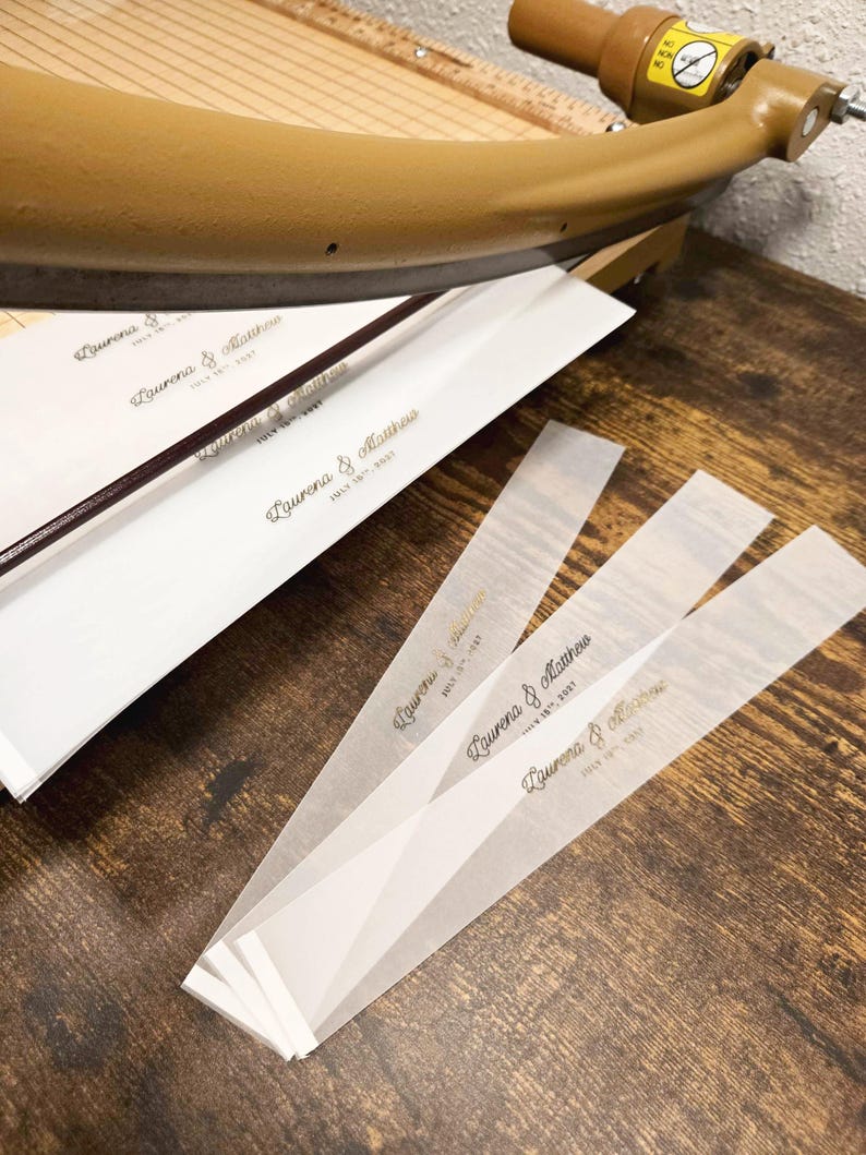 May include: Close-up of wedding invitation strips with the names "Lauren & Matthew" and the date "July 15th, 2023" printed in gold. The strips are on a wooden surface, with a paper cutter in the background.
