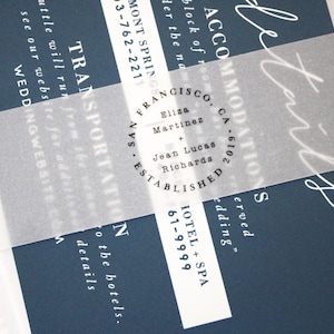 May include: Navy blue wedding invitation with white script and text. A translucent vellum band wraps around the invitation, featuring the names Eliza Martinez and Jean Lucas Richards. The invitation includes details for accommodations and transportation.