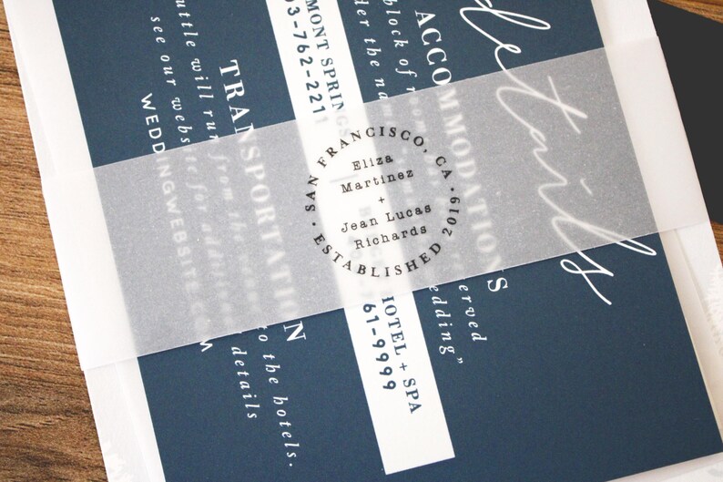 May include: Navy blue wedding invitation with white script and text. A translucent vellum band wraps around the invitation, featuring the names Eliza Martinez and Jean Lucas Richards. The invitation includes details for accommodations and transportation.