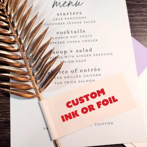 May include: A white menu card with black text listing starters, cocktails, soup, salad, and entrees. A metallic gold palm leaf decoration is placed over the menu. A white paper strip with the red text CUSTOM INK OR FOIL is attached to the leaf.