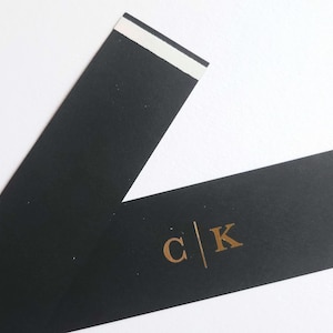 May include: Two black paper strips with gold lettering and a white adhesive strip. The gold lettering includes the initials "C" and "K" separated by a vertical line. The strips are on a white background.