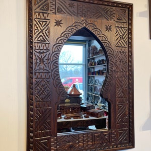 May include: A dark brown wooden mirror with intricate carved designs. The mirror has a traditional Moroccan arch shape and is framed by a thick, ornate border.