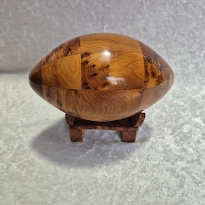 May include: A wooden football shaped ornament made of multiple pieces of wood with a natural wood finish. The ornament is displayed on a small wooden stand.