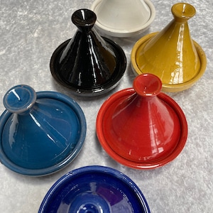 Könnte beinhalten: Sechs Keramik-Tagine in verschiedenen Farben, darunter Weiß, Schwarz, Gelb, Blau und Rot. Die Tagine sind in einem Kreis auf einer weißen Oberfläche angeordnet.