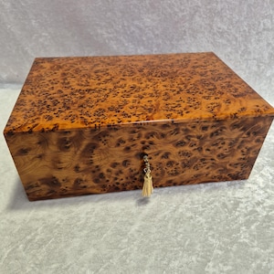 May include: A rectangular wooden box with a glossy finish. The box is made of burl wood, featuring a unique pattern of dark brown spots on a lighter brown background. A small gold-coloured tassel hangs from the keyhole.