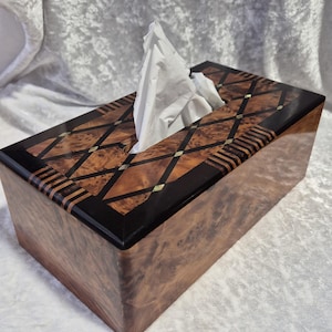 May include: A rectangular wooden tissue box with a decorative inlaid top. The top features a geometric pattern of dark and light wood, with black borders. White tissues are visible. The box is made of a dark wood.