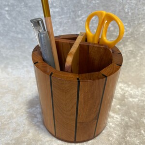 May include: A wooden desk organizer with multiple compartments. It contains a pen, pencil, and yellow scissors. The organizer is made of light brown wood with black accent lines. The organizer is cylindrical in shape.