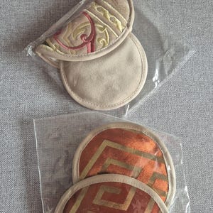 Peut inclure: Deux ensembles de dessous de verre décoratifs dans un emballage plastique transparent. Un ensemble présente un motif beige brodé avec des accents rouges et dorés. L'autre ensemble a un motif géométrique orange avec des détails dorés.