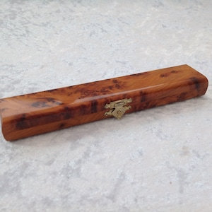 Op de afbeelding: Een rechthoekige houten kist met een glanzende afwerking en een decoratieve messing sluiting. De kist is gemaakt van een warm getint hout met een uniek wortelnerfpatroon, wat duidt op een handgemaakt item.