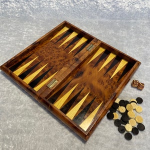 May include: A wooden backgammon board with a burl wood finish. The board is open and the playing surface is visible. There are black and white game pieces and dice on the table next to the board.
